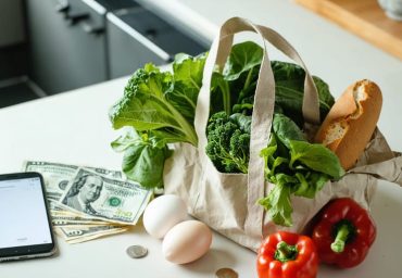 Reusable grocery bag filled with fresh produce next to Canadian banknotes, coins, and a smartphone on a kitchen countertop, with a modern kitchen softly blurred in the background.